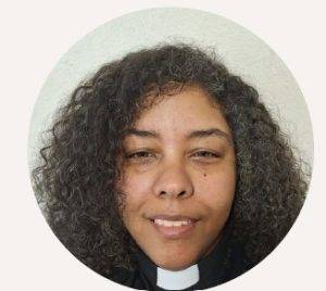 Photo is of a multiracial woman smilingwith long curly natural hair wearing ablack clerical shirt with white tab collar